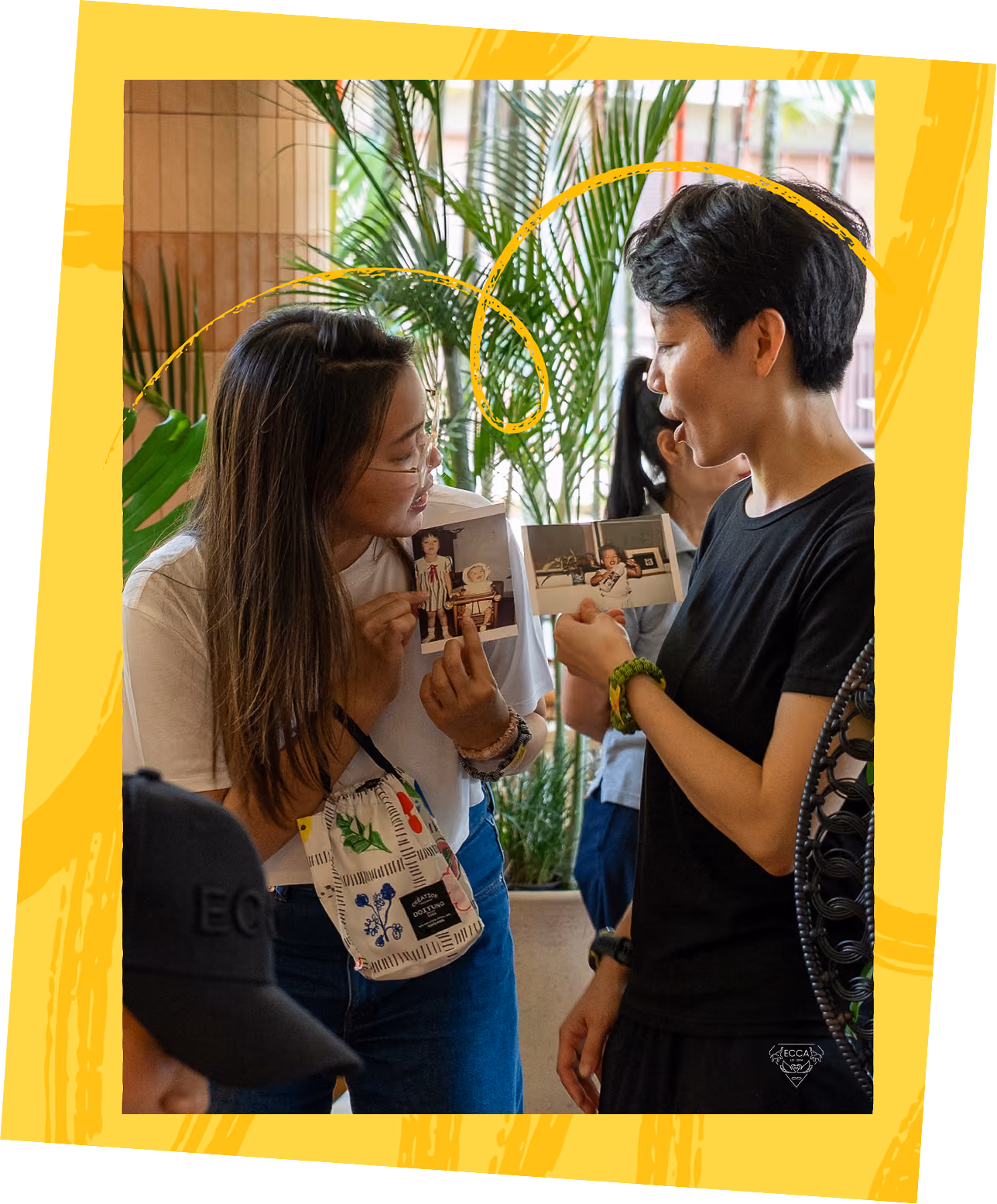Two people in conversation at an indoor gathering, one holding a printed photograph, surrounded by palm plants.