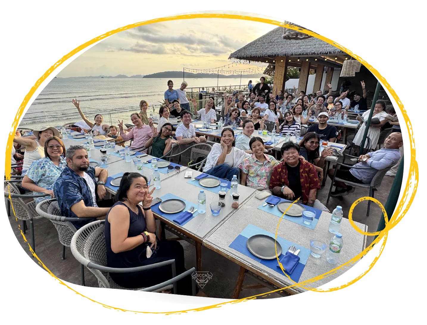 Partners seated along a long table beside the sea at dusk, waving at the camera.