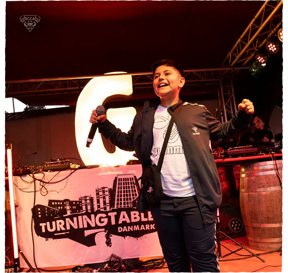 A young performer holding a microphone on stage in front of a Turning Tables Denmark banner.